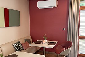 Essbereich mit zwei weißen Tischen, einer beige gepolsterten Eckbank mit Kissen und einem roten Stuhl vor einer rot gestrichenen Wand mit Klimagerät und Fenster mit grauem Vorhang