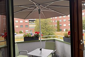 Balkon mit quadratischem Tisch, zwei Stühlen, großem Sonnenschirm und Blumenkästen vor einem mehrstöckigen Wohngebäude