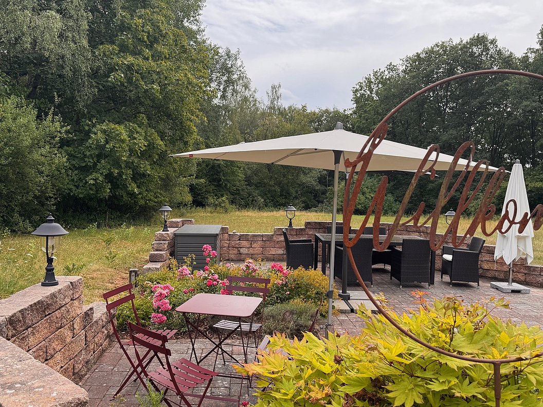 Terrasse mit Tischen, Stühlen, Sonnenschirmen und Pflanzen vor einem Wald