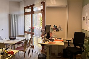 Büroecke mit Schreibtisch, Computer, Bürostuhl, mehreren Stühlen um einen Tisch und großer Fensterfront mit Blick nach draußen