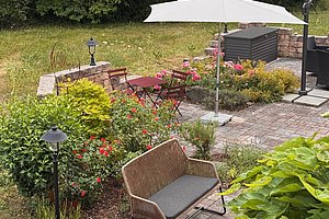 Garten mit gepflasterter Terrasse, Sitzgruppe mit Tisch und Stühlen, Sonnenschirm und Pflanzenbeeten