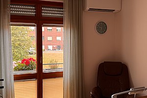 Fenster mit halb geöffnetem Plissee, dahinter Blumenkasten mit roten Blumen, daneben brauner Sessel und runde Wanduhr mit schwarzem Zifferblatt